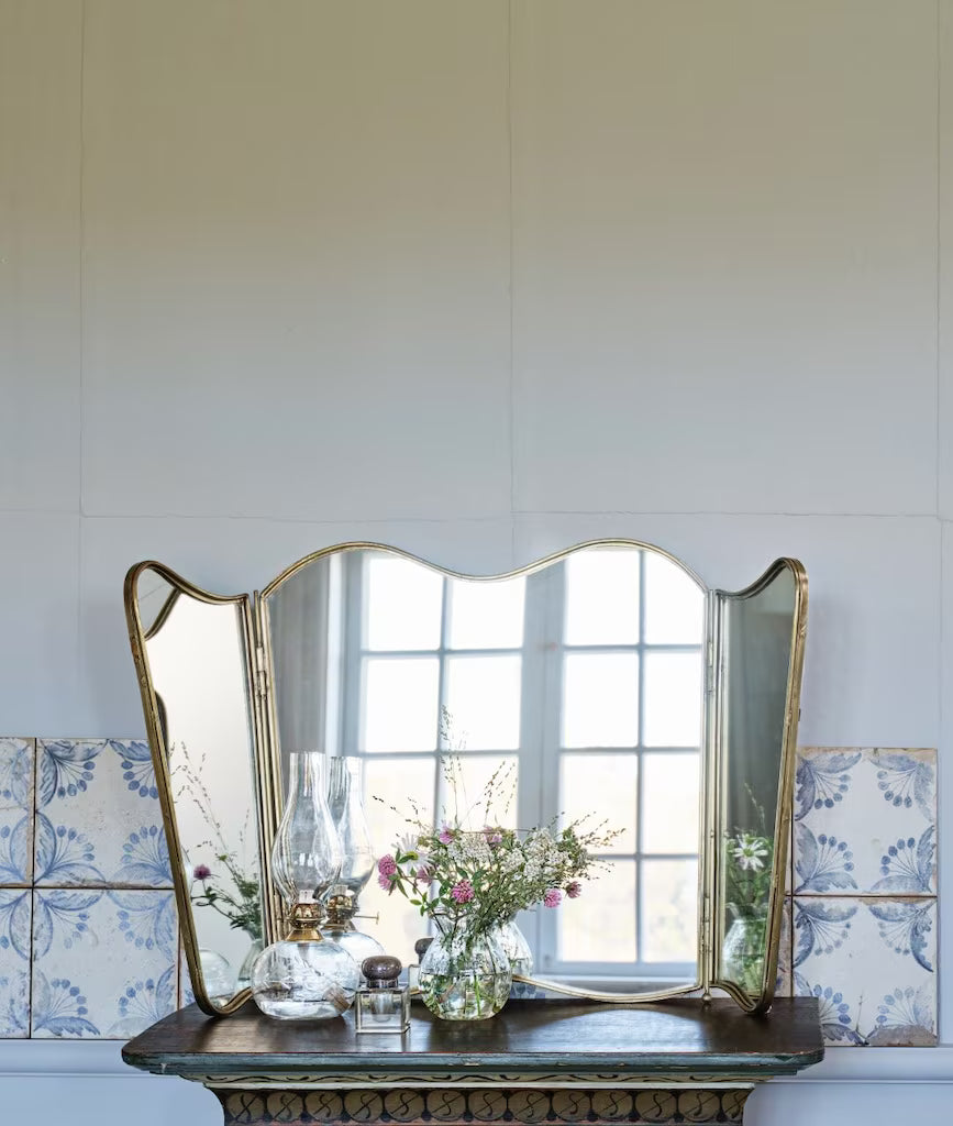 Espejo de tres hojas con acabado en latón envejecido sobre cómoda con flores y lámparas de aceite. Estilo vintage elegante y artesanal.